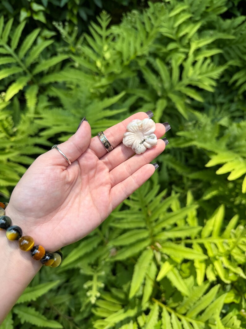 May include: A carved, off-white flower held in a hand, set against a backdrop of green foliage. The flower has detailed petals. A bracelet with yellow and black beads is visible on the wrist, along with rings on the fingers.