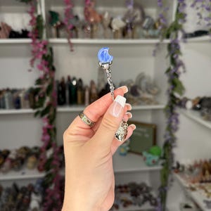May include: A silver pendant with a blue crescent moon and a decorative charm. The pendant is held up in front of a shelf filled with various items, with purple flowers in the background.