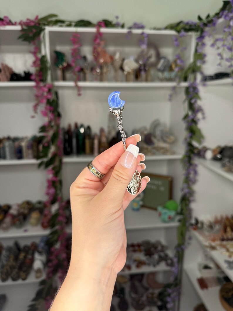 May include: A silver pendant with a blue crescent moon and a decorative charm. The pendant is held up in front of a shelf filled with various items, with purple flowers in the background.