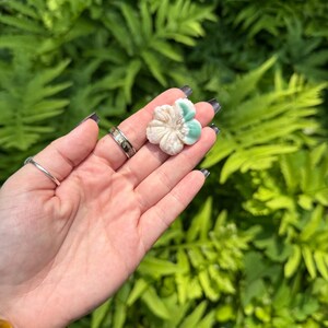 May include: A carved flower-shaped stone with a cream and teal color scheme. The flower is held in a hand against a backdrop of green foliage. The stone appears to be a decorative item or a piece of jewelry.