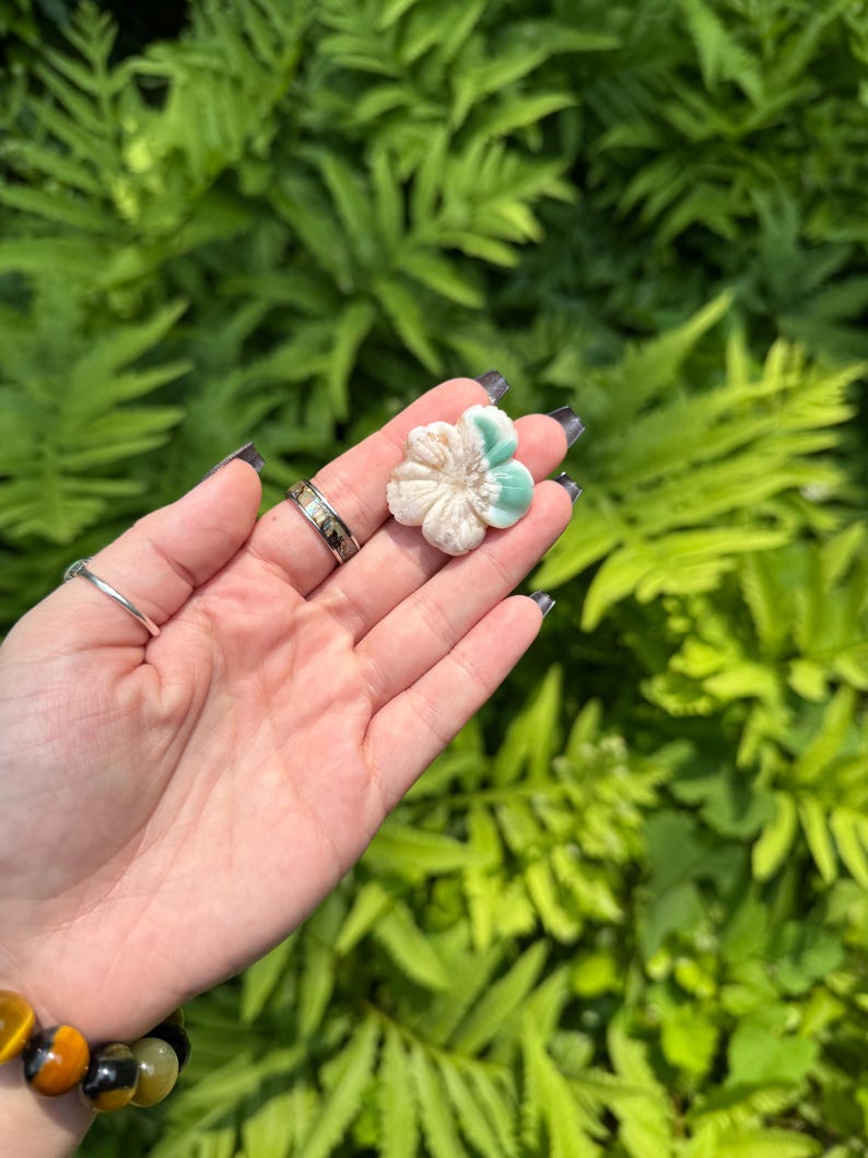 May include: A carved flower-shaped stone with a cream and teal color scheme. The flower is held in a hand against a backdrop of green foliage. The stone appears to be a decorative item or a piece of jewelry.