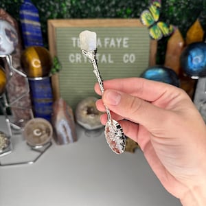 May include: A silver crystal spoon with a floral design on the bowl and handle, topped with a white crystal. The background includes a sign that reads "TEA FAYE CRYSTAL CO."