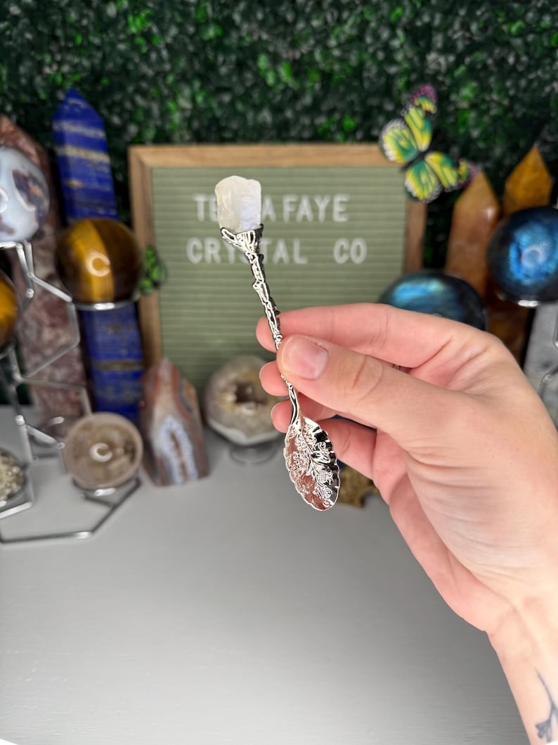 May include: A silver crystal spoon with a floral design on the bowl and handle, topped with a white crystal. The background includes a sign that reads "TEA FAYE CRYSTAL CO."