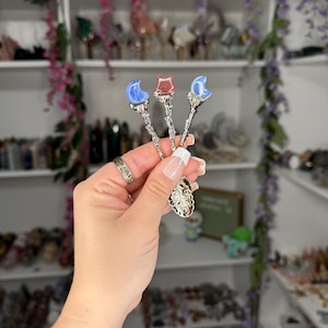 May include: Three decorative spoons with celestial designs. Each spoon features a different gemstone: a blue crescent moon, a red star, and a blue crescent moon. The spoons have ornate silver handles and are held in a hand.