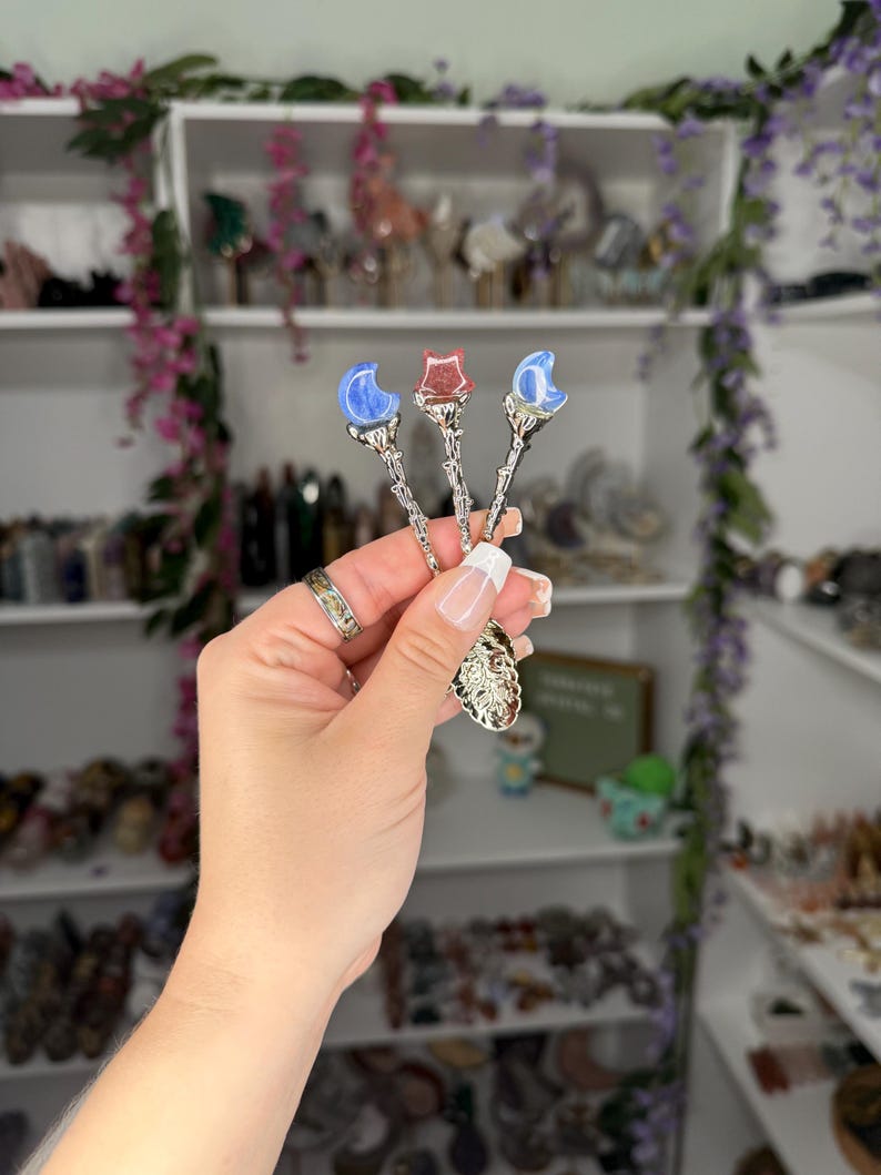May include: Three decorative spoons with celestial designs. Each spoon features a different gemstone: a blue crescent moon, a red star, and a blue crescent moon. The spoons have ornate silver handles and are held in a hand.