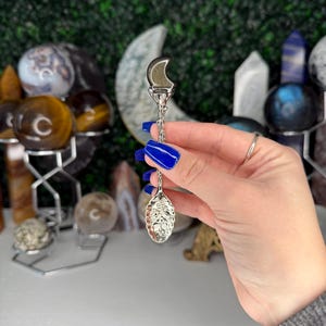 May include: A silver tea infuser featuring a crescent moon and leaf-shaped basket. The infuser is held by a person with blue nail polish. The background includes various crystals and spheres, creating a mystical aesthetic.