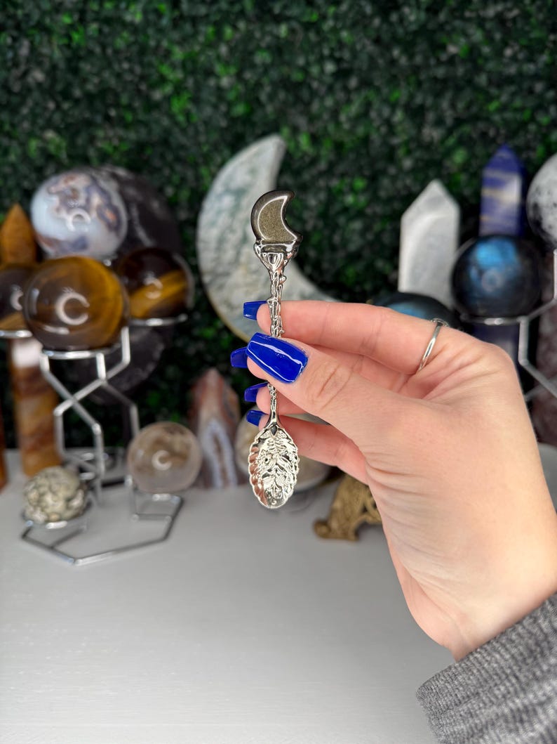 May include: A silver tea infuser featuring a crescent moon and leaf-shaped basket. The infuser is held by a person with blue nail polish. The background includes various crystals and spheres, creating a mystical aesthetic.