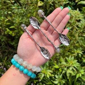 May include: Three silver spoons with leaf-shaped bowls and crystal ball accents are displayed. Two beaded bracelets in shades of blue and white are also visible. The background is a lush green with natural light.
