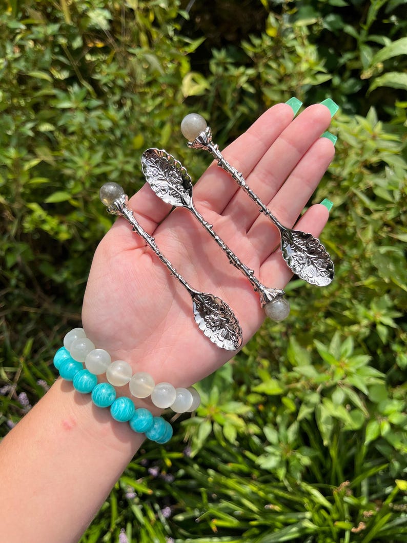 May include: Three silver spoons with leaf-shaped bowls and crystal ball accents are displayed. Two beaded bracelets in shades of blue and white are also visible. The background is a lush green with natural light.