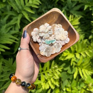 May include: A wooden square bowl filled with carved flower-shaped stones in shades of white, pink, and green. The bowl is held in a hand wearing a ring and a bracelet with orange, yellow, and black beads, set against a backdrop of green foliage.