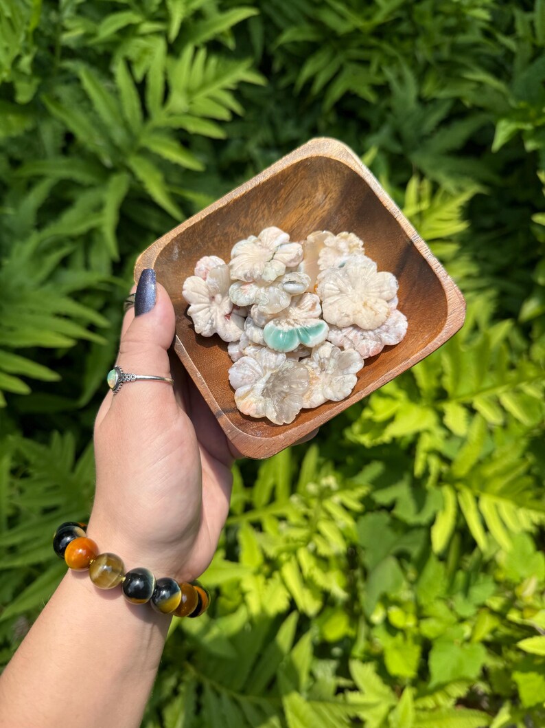 May include: A wooden square bowl filled with carved flower-shaped stones in shades of white, pink, and green. The bowl is held in a hand wearing a ring and a bracelet with orange, yellow, and black beads, set against a backdrop of green foliage.