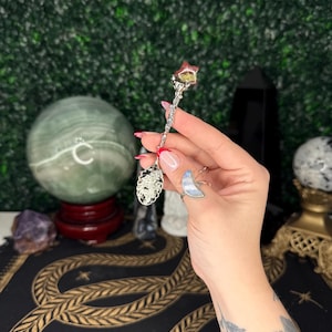 May include: A silver spoon with a decorative handle and a star-shaped crystal. The spoon has an ornate design and is held against a green backdrop. The spoon is likely used for spiritual or decorative purposes.