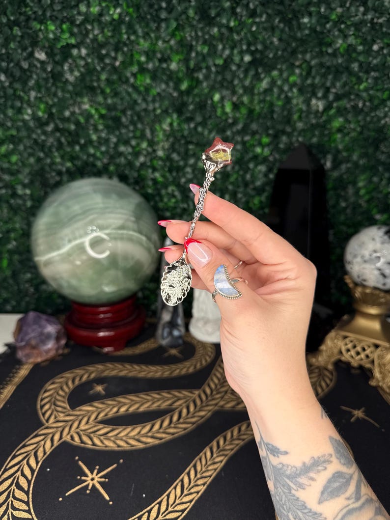 May include: A silver spoon with a decorative handle and a star-shaped crystal. The spoon has an ornate design and is held against a green backdrop. The spoon is likely used for spiritual or decorative purposes.