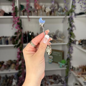 May include: A silver-coloured spoon featuring a blue crescent moon design at the top and a decorative charm at the bottom. The spoon is held in front of a shelf filled with crystals and decorative items.