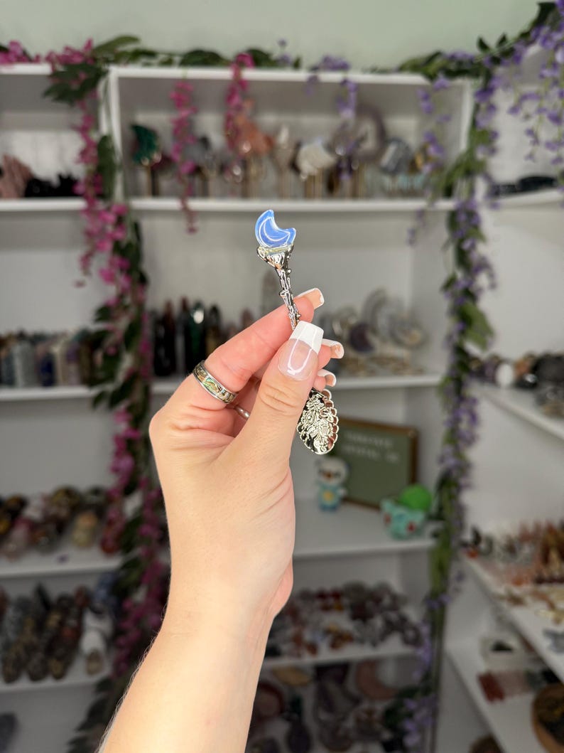 May include: A silver-coloured spoon featuring a blue crescent moon design at the top and a decorative charm at the bottom. The spoon is held in front of a shelf filled with crystals and decorative items.