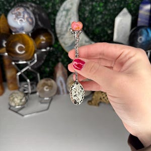 May include: A silver spoon with a decorative handle and a rose-shaped gemstone at the top. The spoon's bowl has an ornate design. The gemstone is pink and orange. The background includes crystal spheres and other decorative items.