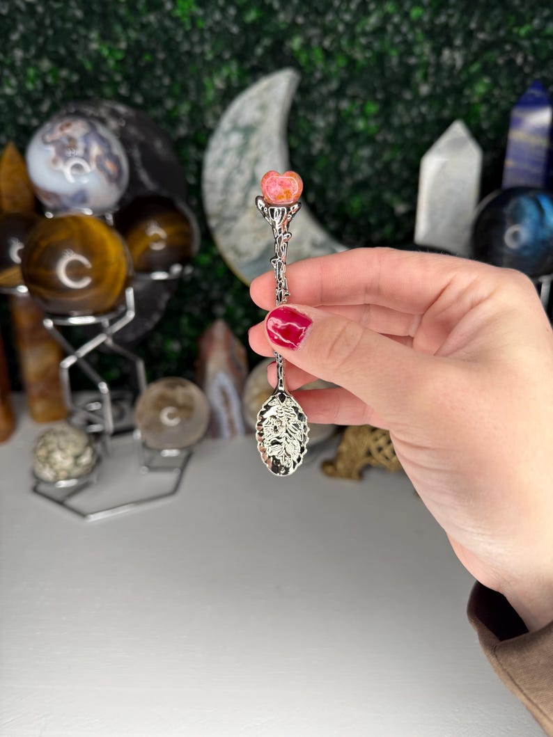 May include: A silver spoon with a decorative handle and a rose-shaped gemstone at the top. The spoon's bowl has an ornate design. The gemstone is pink and orange. The background includes crystal spheres and other decorative items.