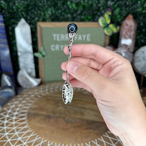 May include: A decorative silver spoon with an ornate handle and a dark blue and black gemstone at the top. The spoon has a detailed, openwork design on the bowl. The text "TERRA FAYE CRY" is visible in the background.