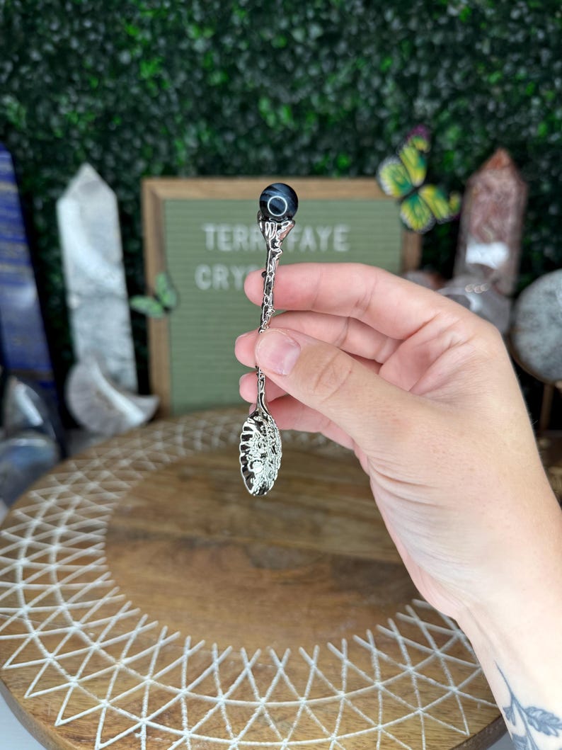 May include: A decorative silver spoon with an ornate handle and a dark blue and black gemstone at the top. The spoon has a detailed, openwork design on the bowl. The text "TERRA FAYE CRY" is visible in the background.