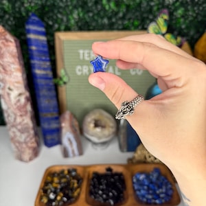 May include: A blue star-shaped lapis lazuli crystal held in a hand. The crystal is small and has a smooth, polished surface.