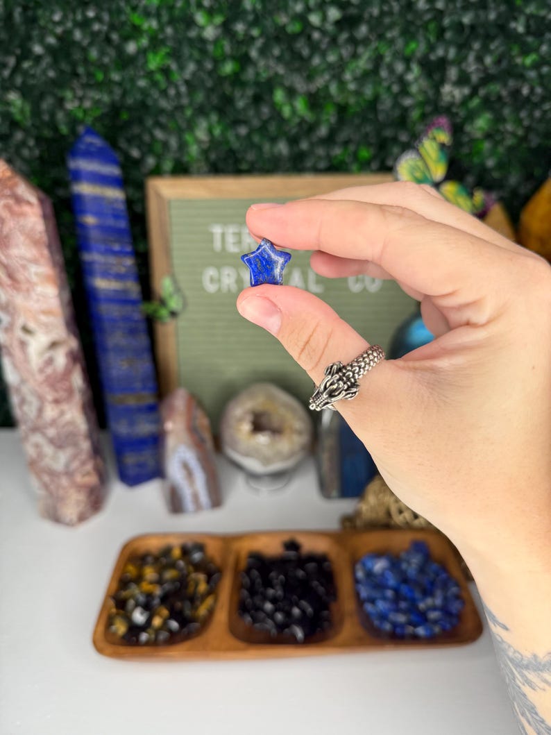 May include: A blue star-shaped lapis lazuli crystal held in a hand. The crystal is small and has a smooth, polished surface.