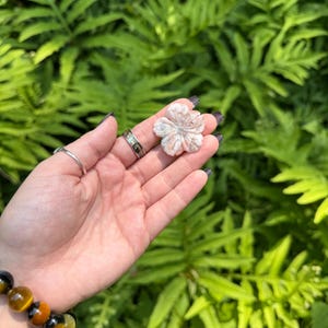 May include: A carved, light pink flower-shaped stone held in a hand. The stone has detailed petal carvings. A bracelet with tiger's eye beads is visible on the wrist. The background is a blurred view of green foliage.