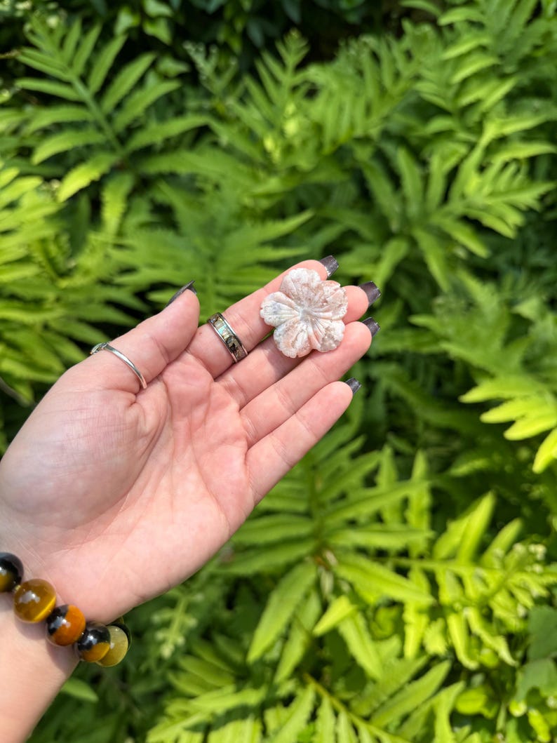 May include: A carved, light pink flower-shaped stone held in a hand. The stone has detailed petal carvings. A bracelet with tiger's eye beads is visible on the wrist. The background is a blurred view of green foliage.
