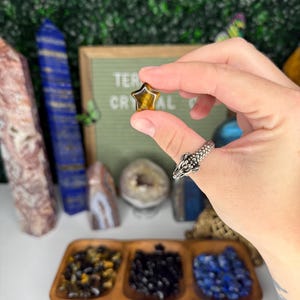 May include: A small, star-shaped tiger's eye gemstone in a hand. The gemstone is a deep golden brown color with a shimmery, iridescent effect. The hand is wearing a silver ring with a dragon design.