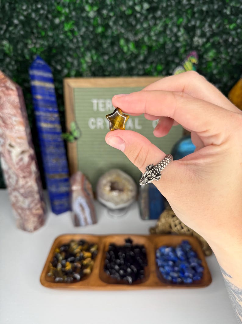 May include: A small, star-shaped tiger's eye gemstone in a hand. The gemstone is a deep golden brown color with a shimmery, iridescent effect. The hand is wearing a silver ring with a dragon design.