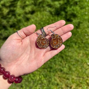 May include: A pair of silver earrings with a brown ammonite fossil design. The earrings are held in a hand with a red beaded bracelet.