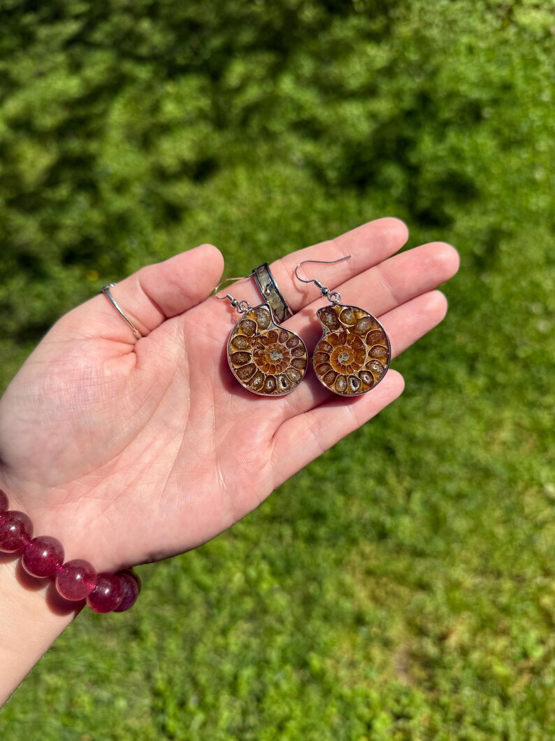 May include: A pair of silver earrings with a brown ammonite fossil design. The earrings are held in a hand with a red beaded bracelet.