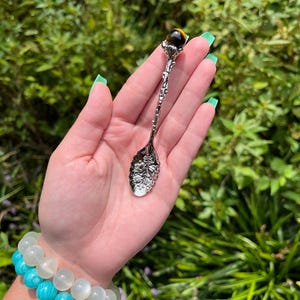 May include: A decorative silver spoon with a floral design and a tiger's eye gemstone at the top. The spoon is held against a backdrop of green foliage. A turquoise and white beaded bracelet is visible.