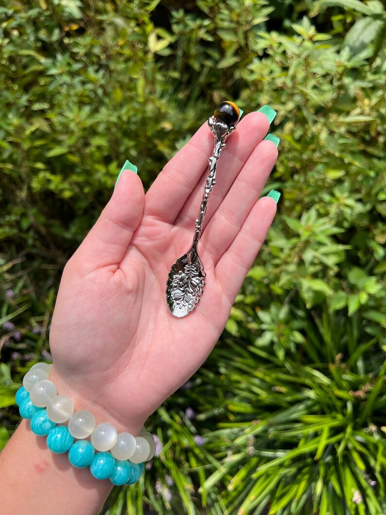 May include: A decorative silver spoon with a floral design and a tiger's eye gemstone at the top. The spoon is held against a backdrop of green foliage. A turquoise and white beaded bracelet is visible.
