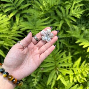 May include: A small, flower-shaped stone carving with a light gray and white color scheme. The carving is held in a hand, with a bracelet of yellow and blue beads on the wrist. The background is a lush green foliage.