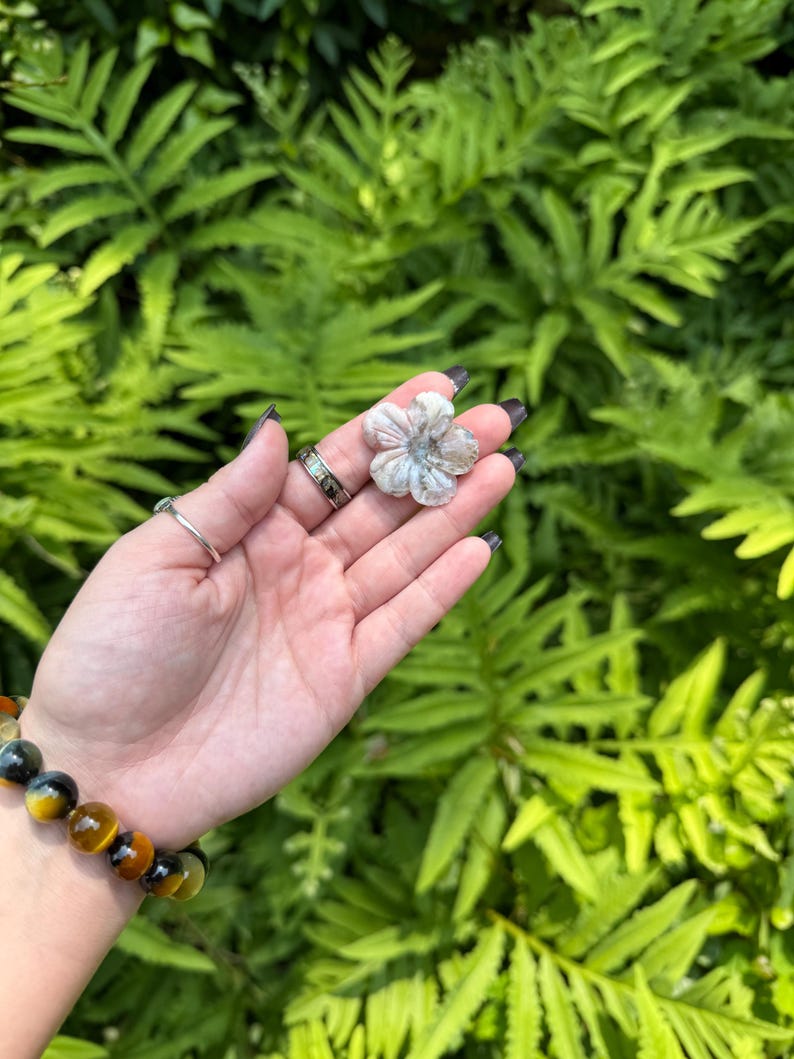 May include: A small, flower-shaped stone carving with a light gray and white color scheme. The carving is held in a hand, with a bracelet of yellow and blue beads on the wrist. The background is a lush green foliage.
