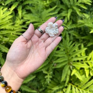 May include: A carved, light-colored stone flower is held in a hand, showcasing intricate details. The flower is the focal point, set against a backdrop of lush green foliage. A bracelet with yellow and brown beads is visible.