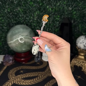 May include: A decorative silver spoon with an amber-coloured crescent moon-shaped stone at the top. The spoon features intricate detailing on the handle and a decorative design at the base. The spoon is held against a backdrop of a green wall and a patterned surface.