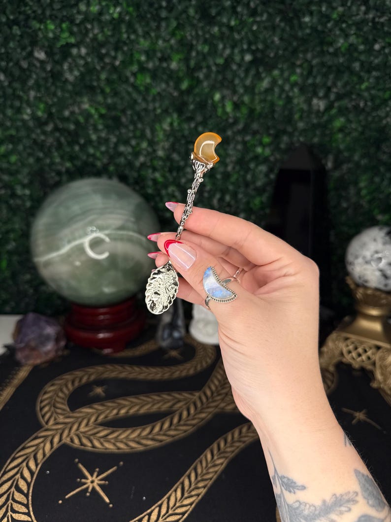 May include: A decorative silver spoon with an amber-coloured crescent moon-shaped stone at the top. The spoon features intricate detailing on the handle and a decorative design at the base. The spoon is held against a backdrop of a green wall and a patterned surface.