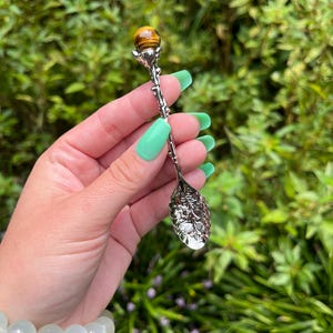 May include: An ornate silver-toned spoon with a tiger's eye gemstone at the top. The spoon has detailed floral and vine designs. The gemstone is a warm brown colour. The spoon is held against a backdrop of green foliage.