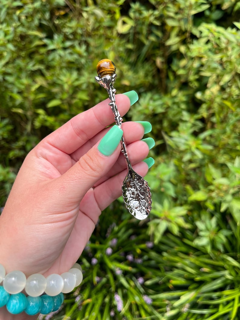 May include: An ornate silver-toned spoon with a tiger's eye gemstone at the top. The spoon has detailed floral and vine designs. The gemstone is a warm brown colour. The spoon is held against a backdrop of green foliage.