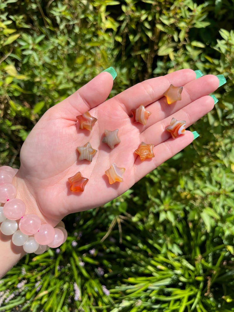 May include: A collection of eight agate star-shaped stones in shades of brown, orange, and white. The stones are arranged on a hand.