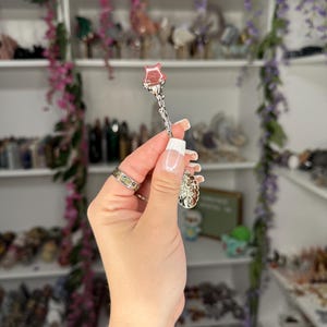 May include: A silver-coloured spoon with a rose-shaped head. The spoon is held up in front of a shelf filled with various items. The rose is a light pink colour.