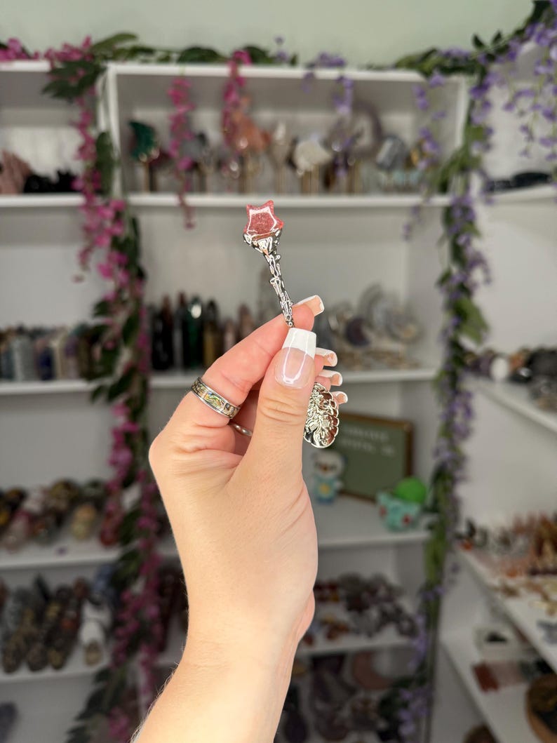 May include: A silver-coloured spoon with a rose-shaped head. The spoon is held up in front of a shelf filled with various items. The rose is a light pink colour.