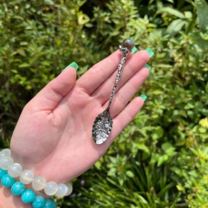 May include: A silver decorative spoon with an intricate handle and a leaf-shaped bowl. The spoon is topped with a small, round, iridescent stone. The background is a lush green garden, with a bracelet of turquoise and white beads.