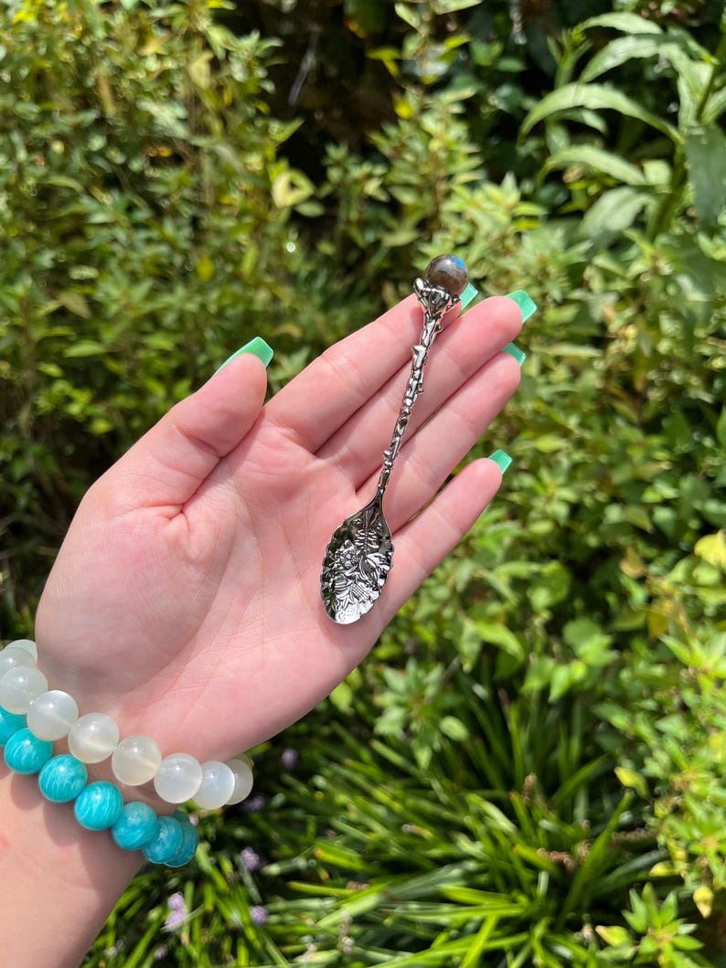 May include: A silver decorative spoon with an intricate handle and a leaf-shaped bowl. The spoon is topped with a small, round, iridescent stone. The background is a lush green garden, with a bracelet of turquoise and white beads.