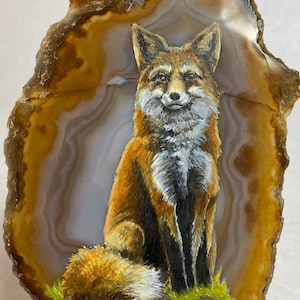May include: A hand-painted red fox on a slice of agate. The fox is sitting with its tail curled around its legs. The agate is a light brown colour with white and brown bands.