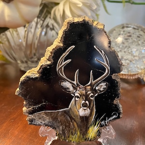 May include: A hand-painted wooden slice featuring a detailed depiction of a white-tailed deer with antlers against a black background. The artwork is signed "M. Morrison" in the bottom right corner.