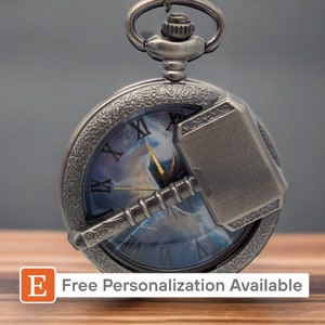 Puede incluir: Reloj de bolsillo plateado con una esfera azul y blanca que representa el martillo de Thor, Mjolnir. El reloj tiene marcadores de hora en números romanos y una cadena plateada.