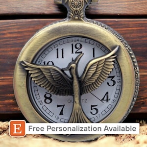 May include: A gold-toned pocket watch with a white face and black Roman numeral hour markers. The watch features a decorative design of a bird with outstretched wings. The watch face also includes the text "QU TZ".