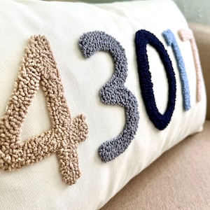 Op de afbeelding: Een wit kussen met de cijfers 4301 geborduurd in beige, grijs, donkerblauw en lichtblauw garen.